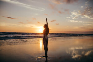 Junge-Frau-steht-mit-weißem-Bikini-am-Strand-und-zeigt-ein-Peace-Zeichen-beim-Sonnenuntergang-buchen1.de